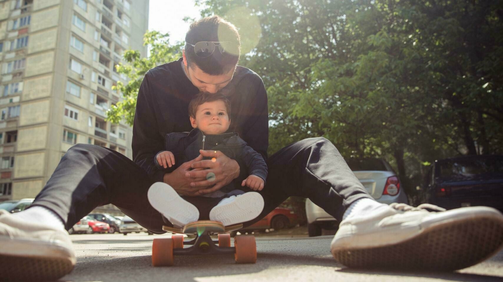 Vater sitz mit einem Kind auf dem Skateboard und hält es mit beiden Händen